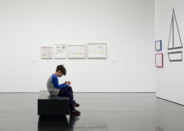 Boy sitting, looking only at his phone in a museum, surrounded by paintings