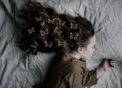 A girl is sleeping, and her brown, curly hair is covered by butterflies as it falls over the pillow 